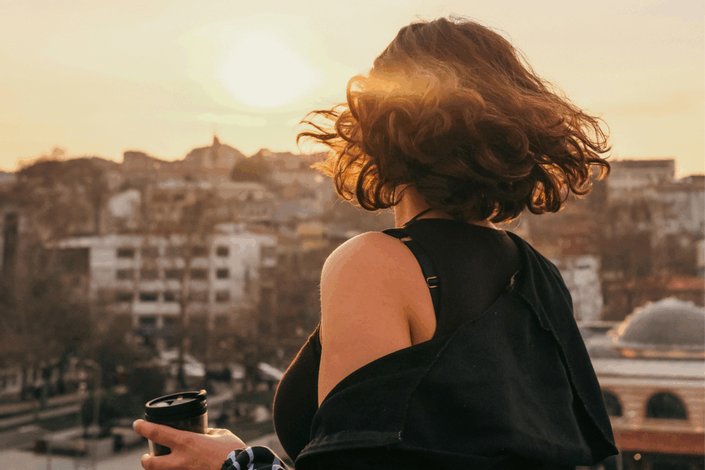 Une femme prend un moment pour elle en savourant son café au lever du soleil, avec la ville en arrière-plan. Une scène qui illustre la beauté du quotidien et la magie des petites habitudes.